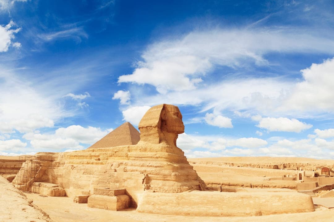 The Great Sphinx of Giza in profile, a pyramid beyond under a cloudy blue sky